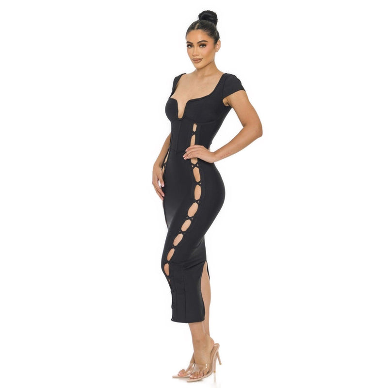 Load image into Gallery viewer, A model poses in a black Bandage Midi Slit Bodycon Dress featuring a unique wired neckline and faux lace-up details along the sides, styled with transparent heels.
