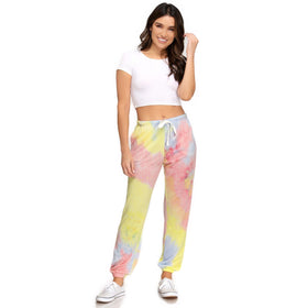 Smiling woman in vibrant yellow and pink tie-dyed jogger pants paired with a white cropped tee, standing against a plain background, showcasing the pants' front view. The comfortable fit and lively colors make a casual yet chic statement.