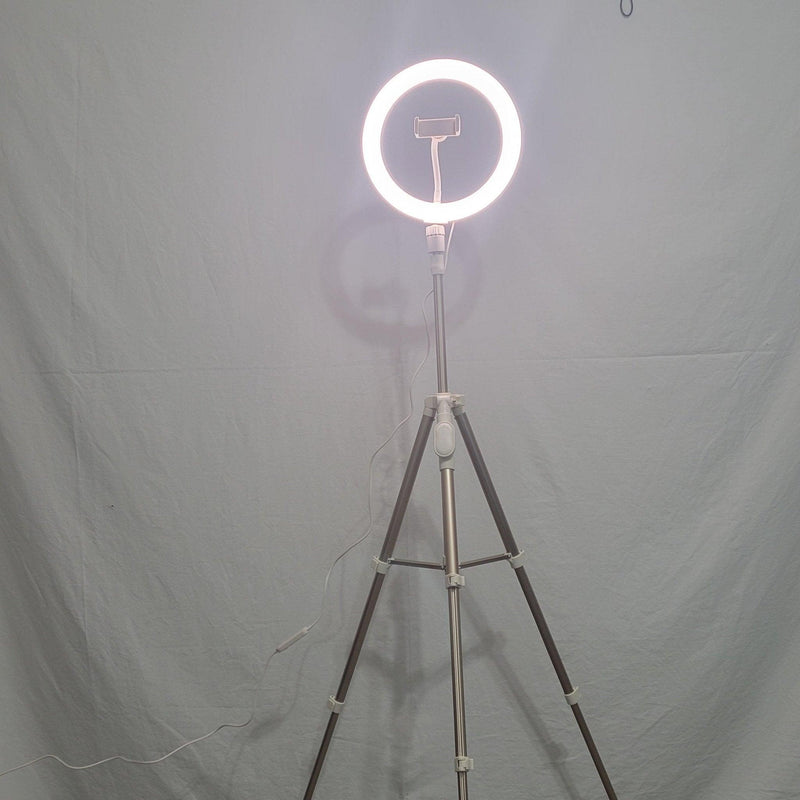Load image into Gallery viewer, A luminous 10-inch Heyday ring light on a tripod, central phone clip empty, casting a soft light in a dimly lit room.
