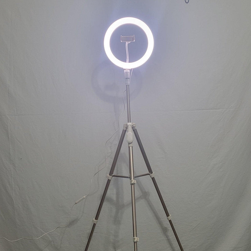 Load image into Gallery viewer, Front view of a 10-inch Heyday ring light mounted on a tripod, illuminated against a grey backdrop, with a power cable trailing to the side.
