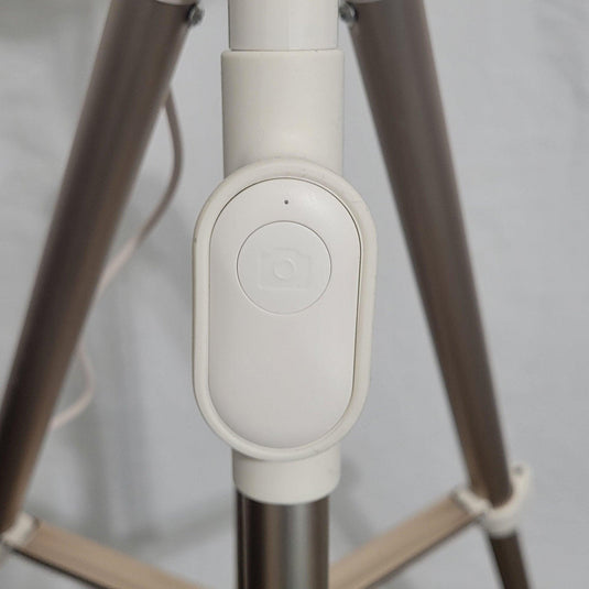 Close-up of the Heyday ring light's remote control with a single power button, set against the tripod's white and rose gold legs.